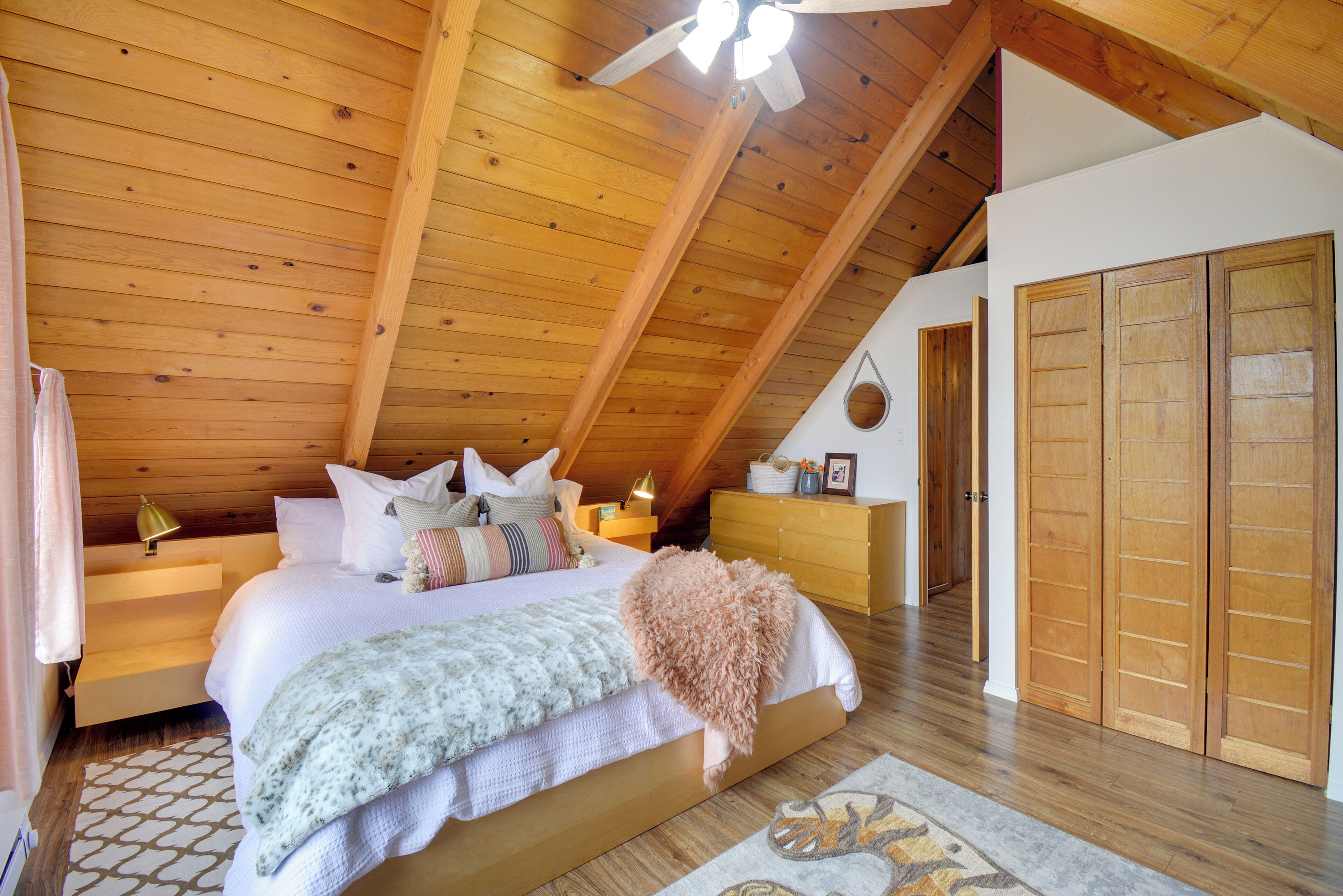 Un dormitorio con techo inclinado, vigas de madera, una cama con almohadas y una colcha, una mesita de noche con lámpara y un armario.
