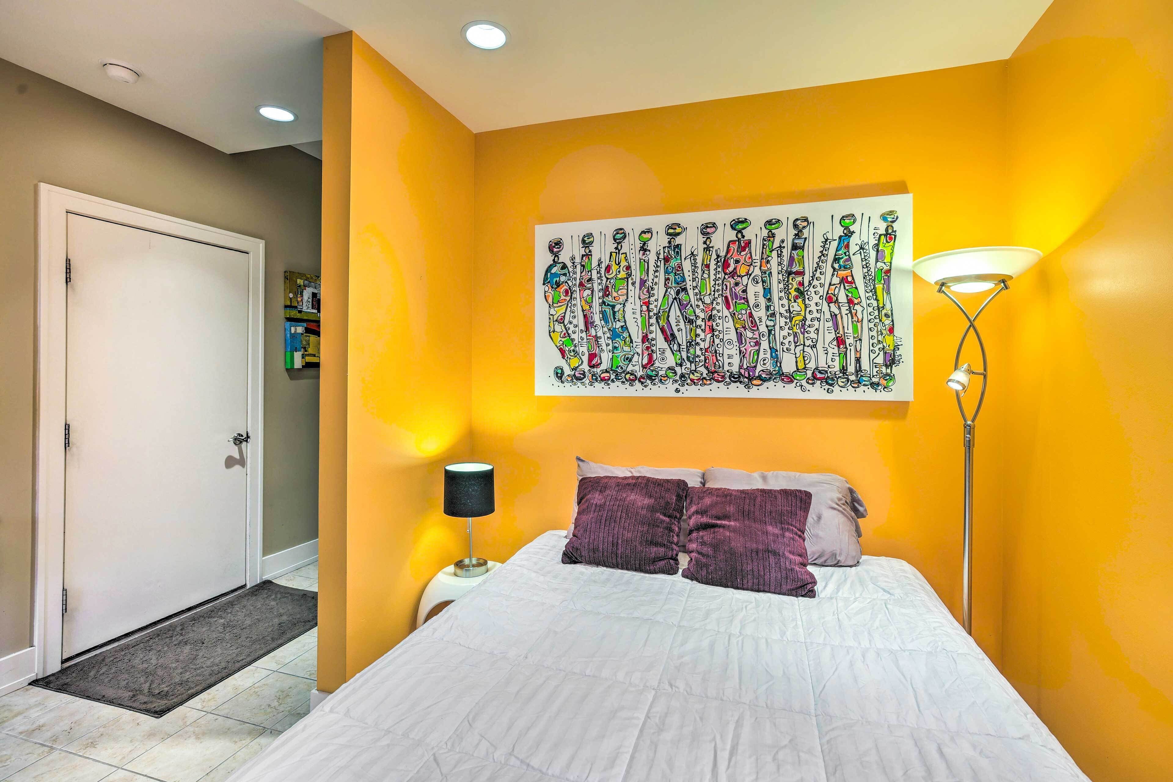 A bedroom with a bed, a nightstand, a lamp, a door, and a wall with a colorful painting.