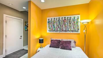 A bedroom with a bed, a nightstand, a lamp, a door, and a wall with a colorful painting.