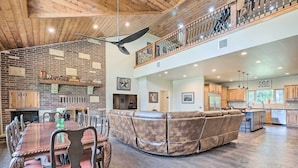 A spacious living area with a fireplace, a high ceiling with a fan, and a kitchen with wooden cabinets.