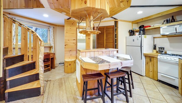 A wooden interior with a central island, bar stools, a kitchen area with appliances, and a staircase. - Unique West Jefferson Home w/ Fire Pit & Grill! (West Jefferson)