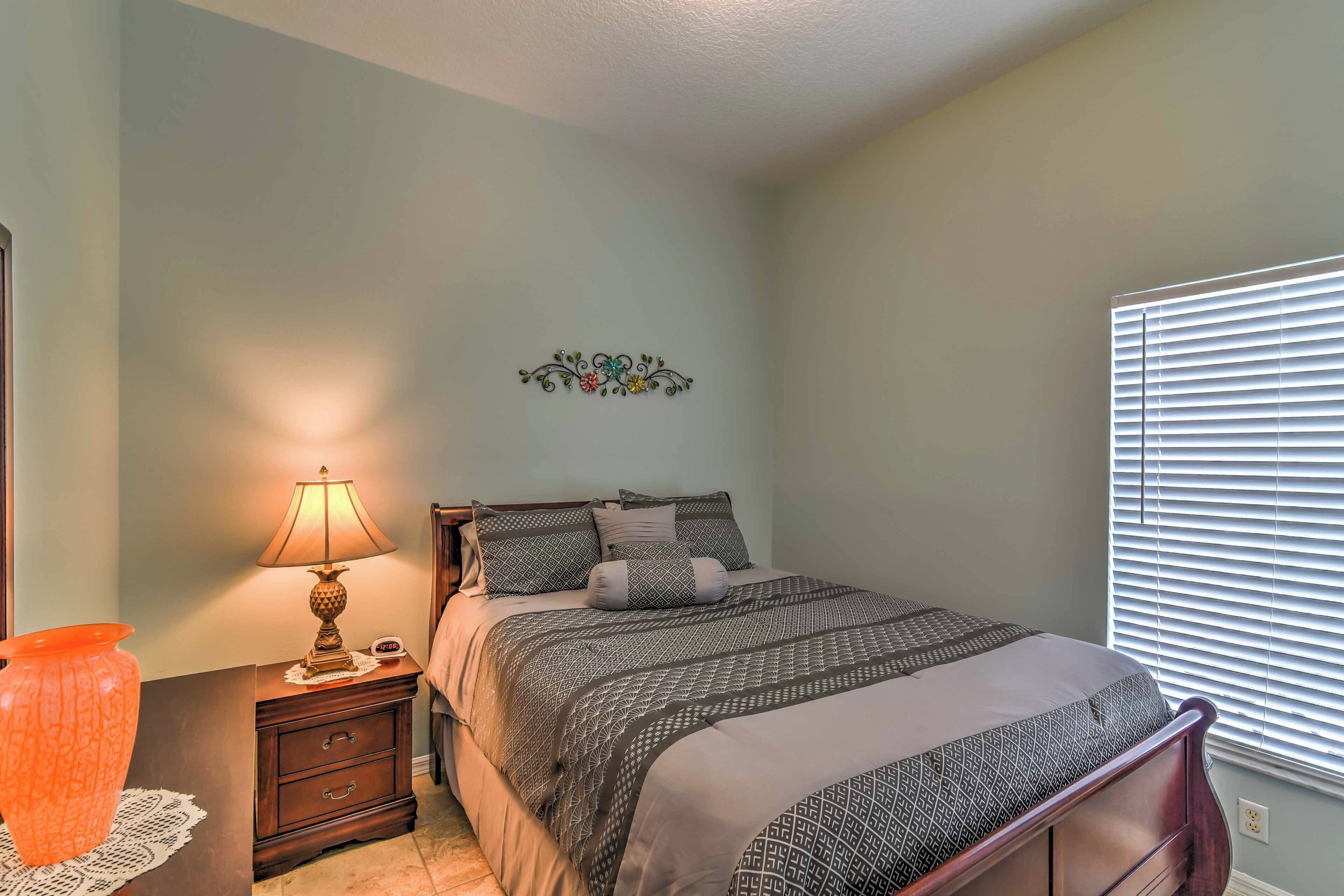 A bedroom with a bed, bedside table, lamp, decorative wall piece, and window with blinds.