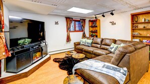 A living room with a large flat-screen TV, leather sofa, and a wooden bookshelf. - Ideally Located Fairbanks Vacation Rental! (Fairbanks)