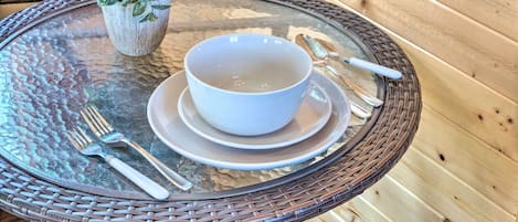 Una mesa de vidrio con un cuenco blanco y cubiertos, una planta en maceta y una silla de mimbre.