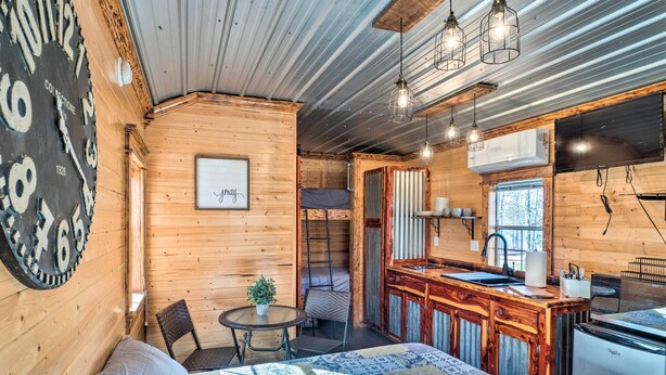 A cozy cabin with a wooden interior, a large clock on the wall, a bed with a patterned blanket, a small dining table, and a kitchen area with a sink and refrigerator. - Sunny 'catfish Cabin' w/ Views of Toledo Bend (Many)