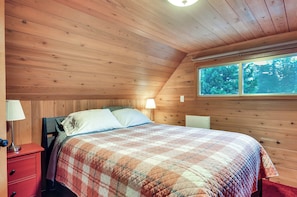 A bedroom with wooden walls, a bed with a plaid quilt, a nightstand with a lamp, and a window with a view of trees.