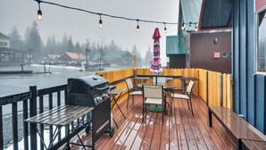 Une terrasse extérieure équipée d’un barbecue, de tables et de chaises, offrant une vue sur le bord de l’eau, avec des bâtiments et des arbres en arrière-plan.