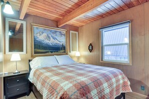 A bedroom with a bed, wooden headboard, two bedside tables with lamps, a large framed painting of a mountain, and a window with blinds.