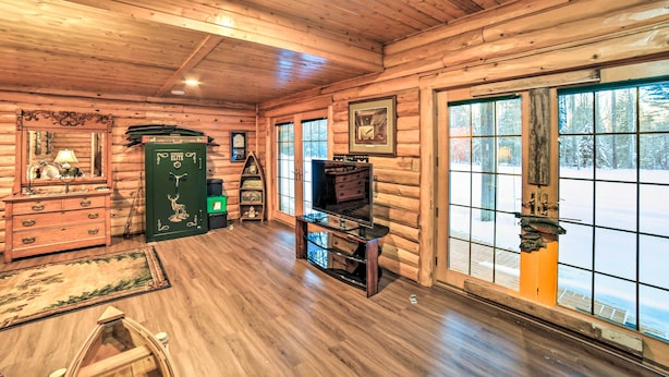 Een vakantiehuisje van binnen, met houten vloeren, een televisie op een meubel, een open haard en grote ramen met uitzicht op sneeuw.