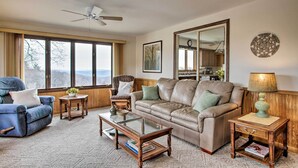 A living room with a leather sofa, a blue armchair, a glass coffee table, a wooden side table with a lamp, and a large mirror on the wall. - Wisconsin River Valley Farmhouse: Fire Pit & View (Portage)