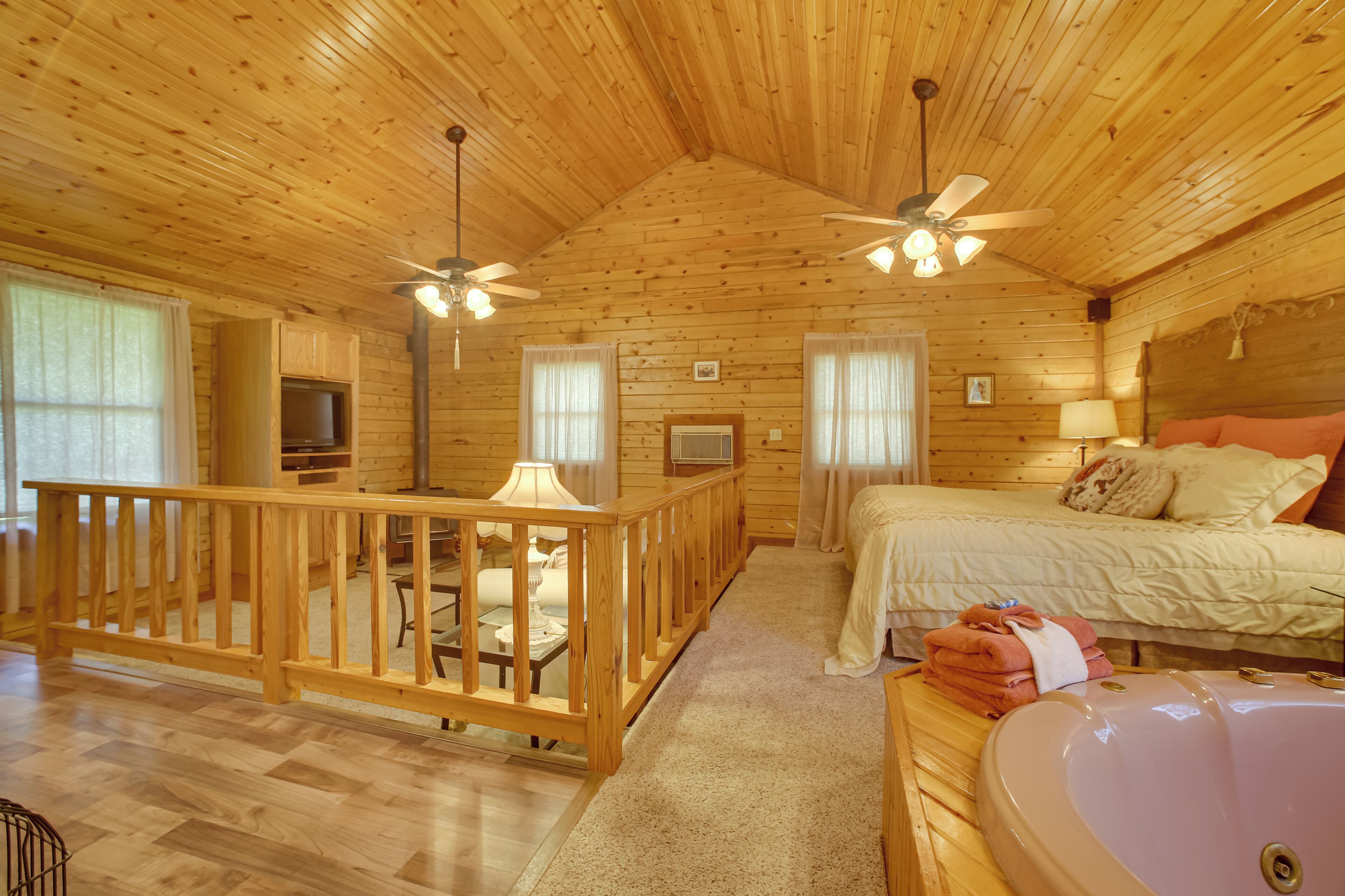 A cabin with a wooden interior, a balcony with a railing, a bed with pillows, a bathroom with a sink, and a ceiling fan.