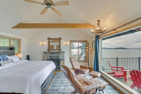 A bedroom with a large bed, a fireplace, a ceiling fan, and a view of a lake. - Cozy Flathead Lake Cabin w/ Picturesque View (Somers)