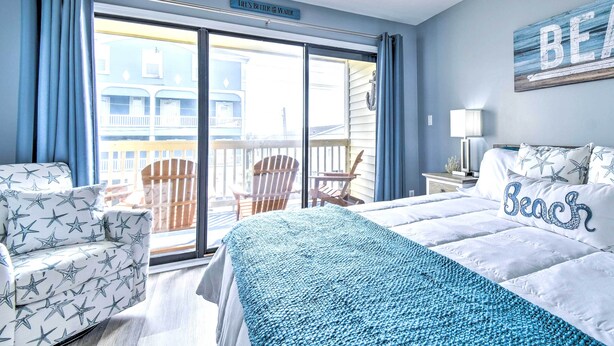 A bedroom with a large bed, a chair, a nightstand, and a view of a balcony.
