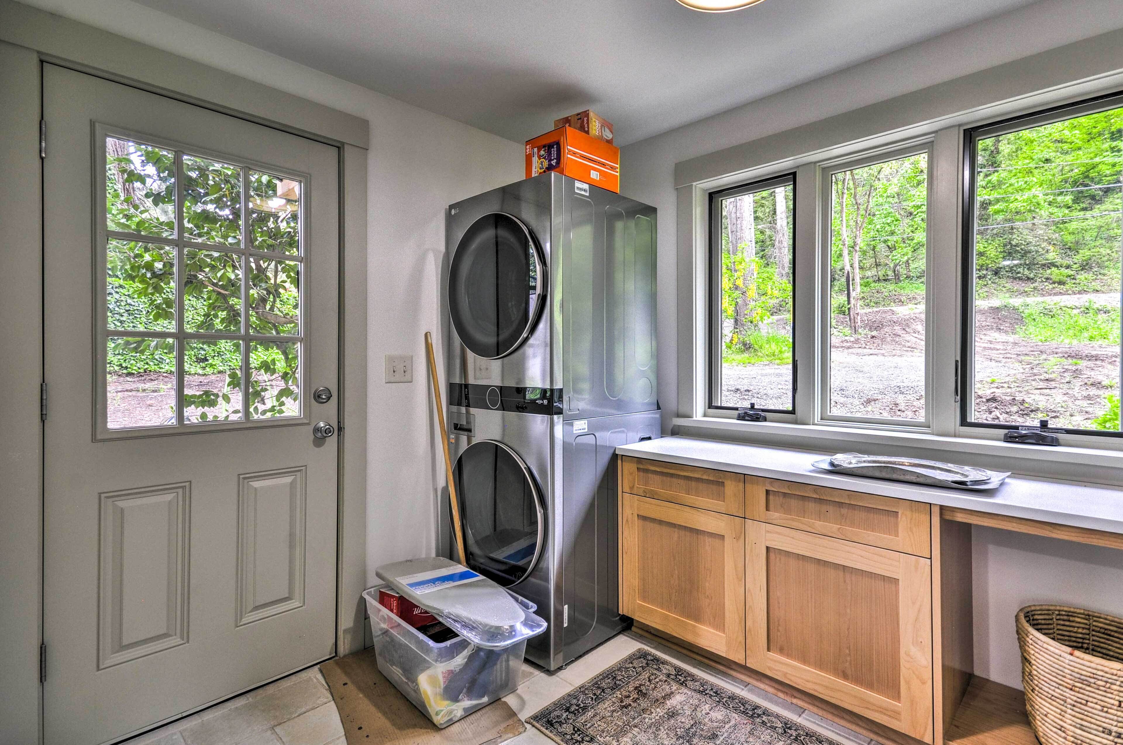 A laundry room with a front-loading washer and dryer, wooden cabinets, a window with a view of greenery, and a door with a window.