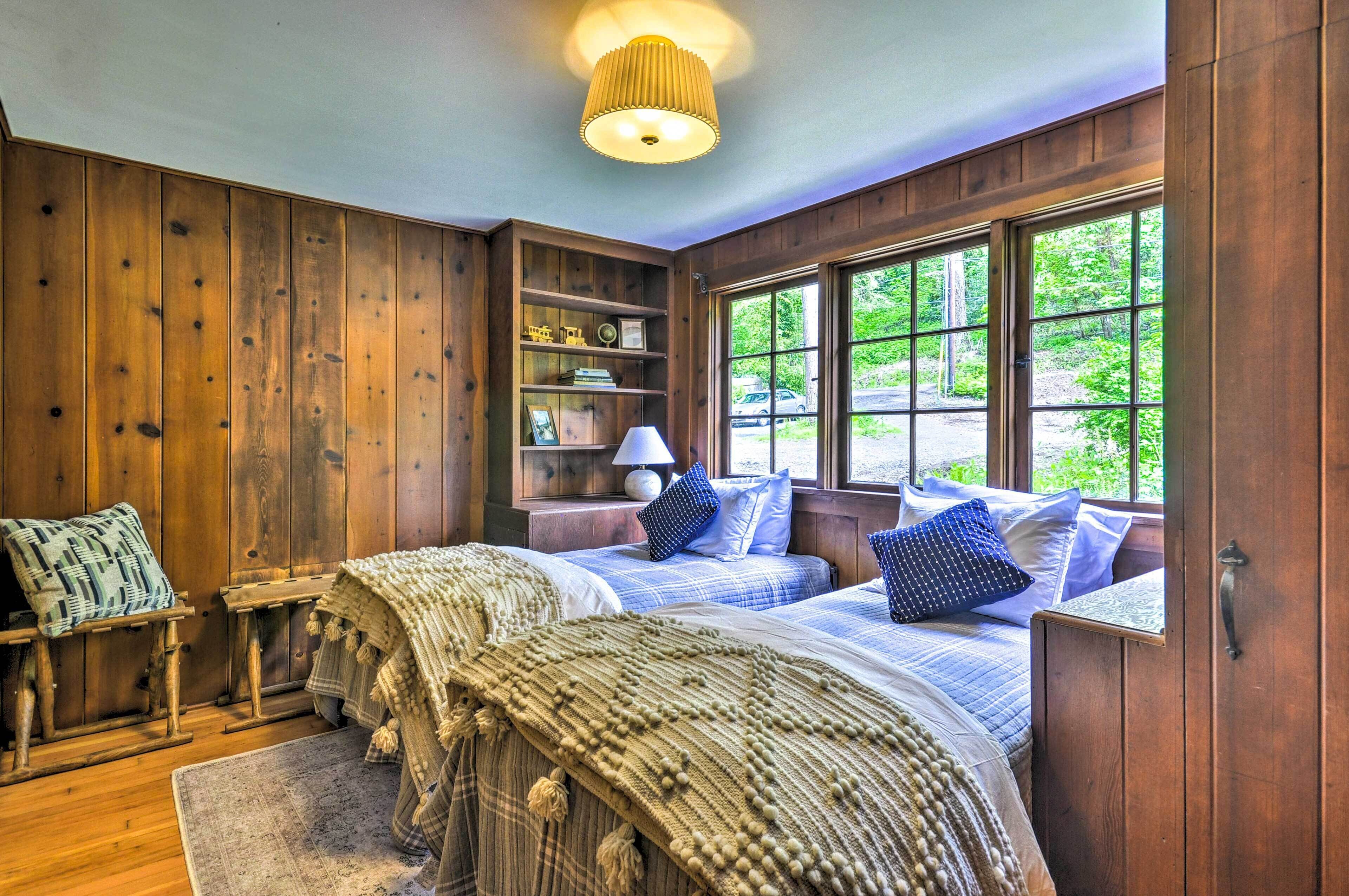 Ein Zimmer aus Holz mit zwei Betten, einem Nachttisch, einer Lampe und einem Fenster mit Blick auf Grünflächen.
