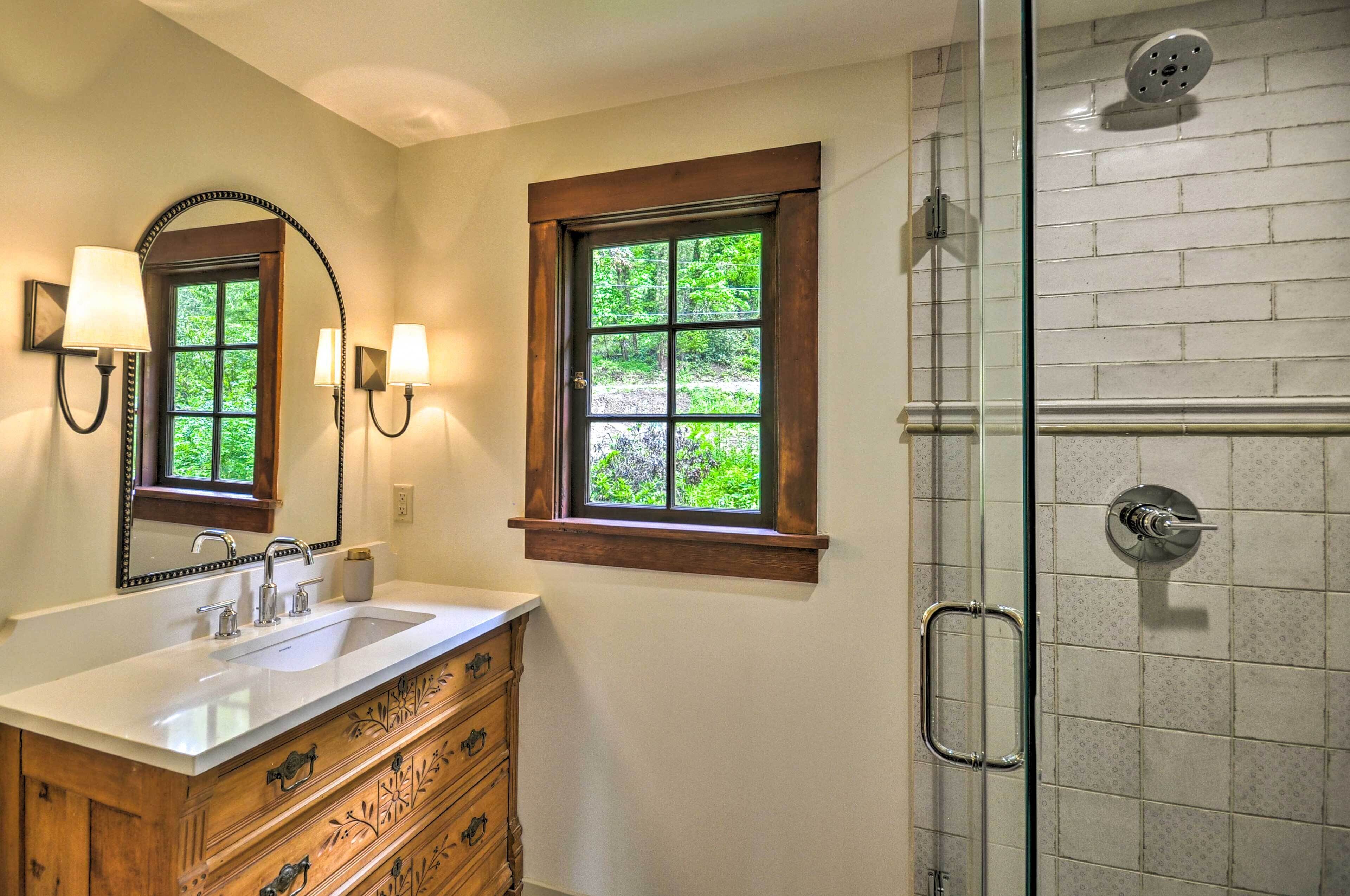 A bathroom with a large mirror, a sink, a vanity with drawers, two windows with wooden frames, and a shower with a glass door.