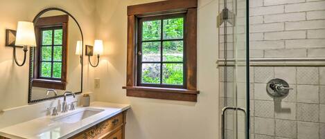 A bathroom with a large mirror, a sink, a vanity with drawers, two windows with wooden frames, and a shower with a glass door.