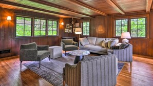 Ein gemütliches Wohnzimmer mit Holzwänden und -böden, großen Fenstern und bequemen Sitzgelegenheiten, darunter eine Couch und Sessel.