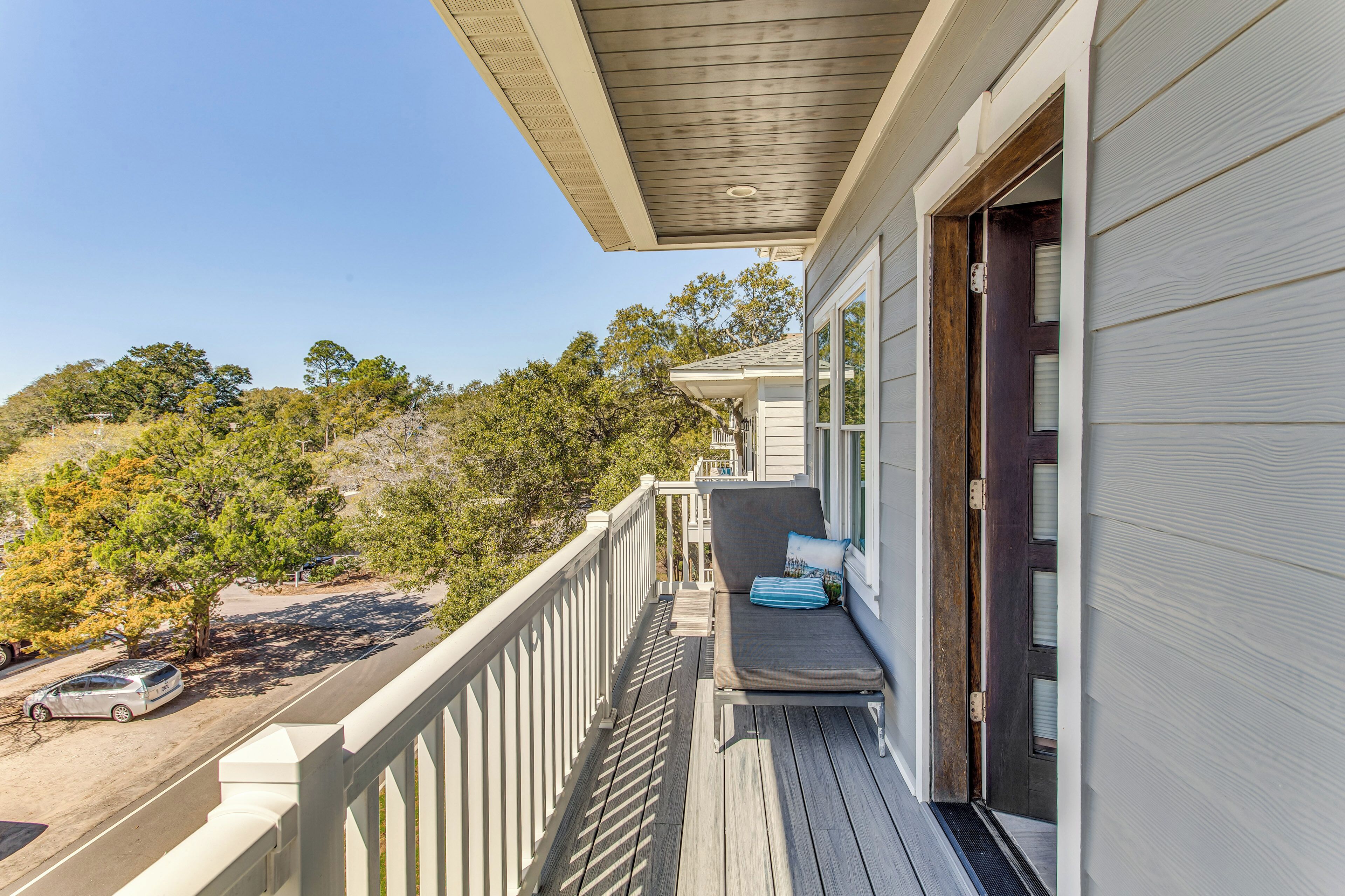 A balcony with a cushioned seating area, a wooden deck, and a view of trees and a road.