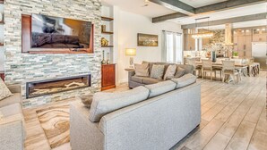 Un salon moderne avec une cheminée en pierre, un grand téléviseur à écran plat, un canapé d’angle gris et un coin repas doté de meubles en bois et d’un lustre.
