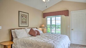 Ein Schlafzimmer mit einem großen Bett, einem Nachttisch, einer Lampe, einem gerahmten Pflanzenmotiv und einem Fenster mit Blick auf Grün.