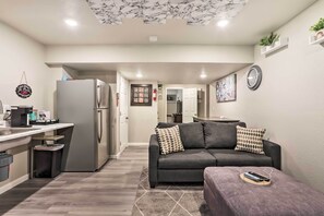 A modern kitchen with a refrigerator, sink, and countertop. There is a gray sofa with patterned cushions in the living area.