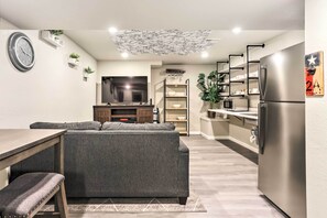 A modern living room with a grey sofa, a wooden coffee table, and a TV. There is a kitchen area with a stainless steel refrigerator and a microwave.