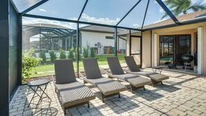 Alpendre coberto com quatro cadeiras de vime e uma pequena mesa, com vista para uma área gramada e um edifício com piscina.