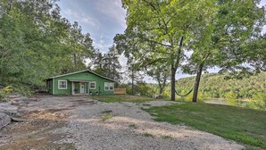 Une maison verte avec une allée en gravier, entourée d’arbres et située au pied d’une colline.