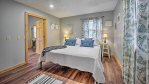 Une chambre à coucher avec un grand lit, un plancher en bois, une porte donnant sur une autre pièce et une fenêtre ornée de rideaux à motifs.