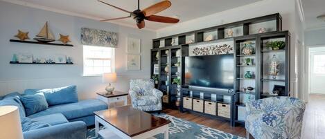 A living room with a blue sofa, matching armchairs, a wooden coffee table, a ceiling fan, and a large TV. Shelves with decorative items line the walls.
