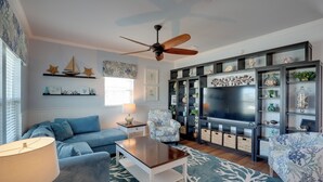 Ein Wohnzimmer mit einem blauen Sofa, einem Holztisch, einem gemusterten Sessel, einem Deckenventilator und einem Fernseher auf einer schwarzen Unterhaltungsanlage.