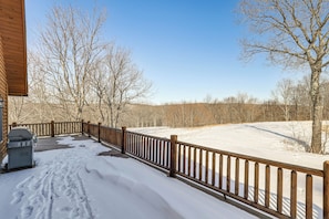 A wooden deck with a snow-covered ground, a barbecue grill, and leafless trees in the background.