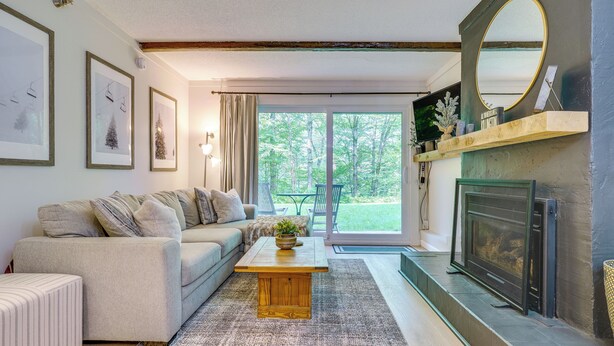 A living room with a sofa, a coffee table, a fireplace, and a large window with a view of trees. - Ski-home Trail: Updated Killington Condo (Killington)
