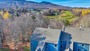 A large blue building with multiple windows and solar panels, surrounded by trees and a golf course in the background. - Ski-home Trail: Updated Killington Condo (Killington)