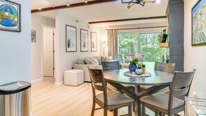 A dining area with a round table, four chairs, and a vase of flowers. There is a sofa and a window with a view of trees. - Ski-home Trail: Updated Killington Condo (Killington)