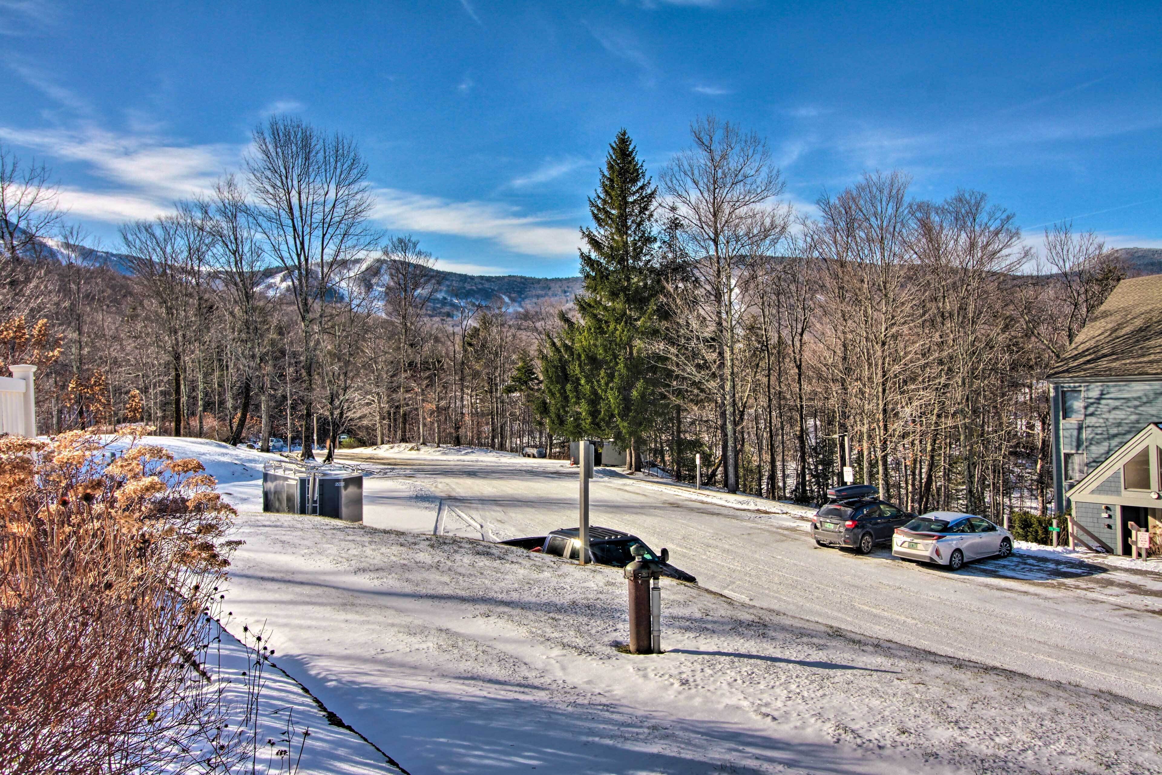 A snowy street with cars parked on the side, a utility box, and a mountain in the background.