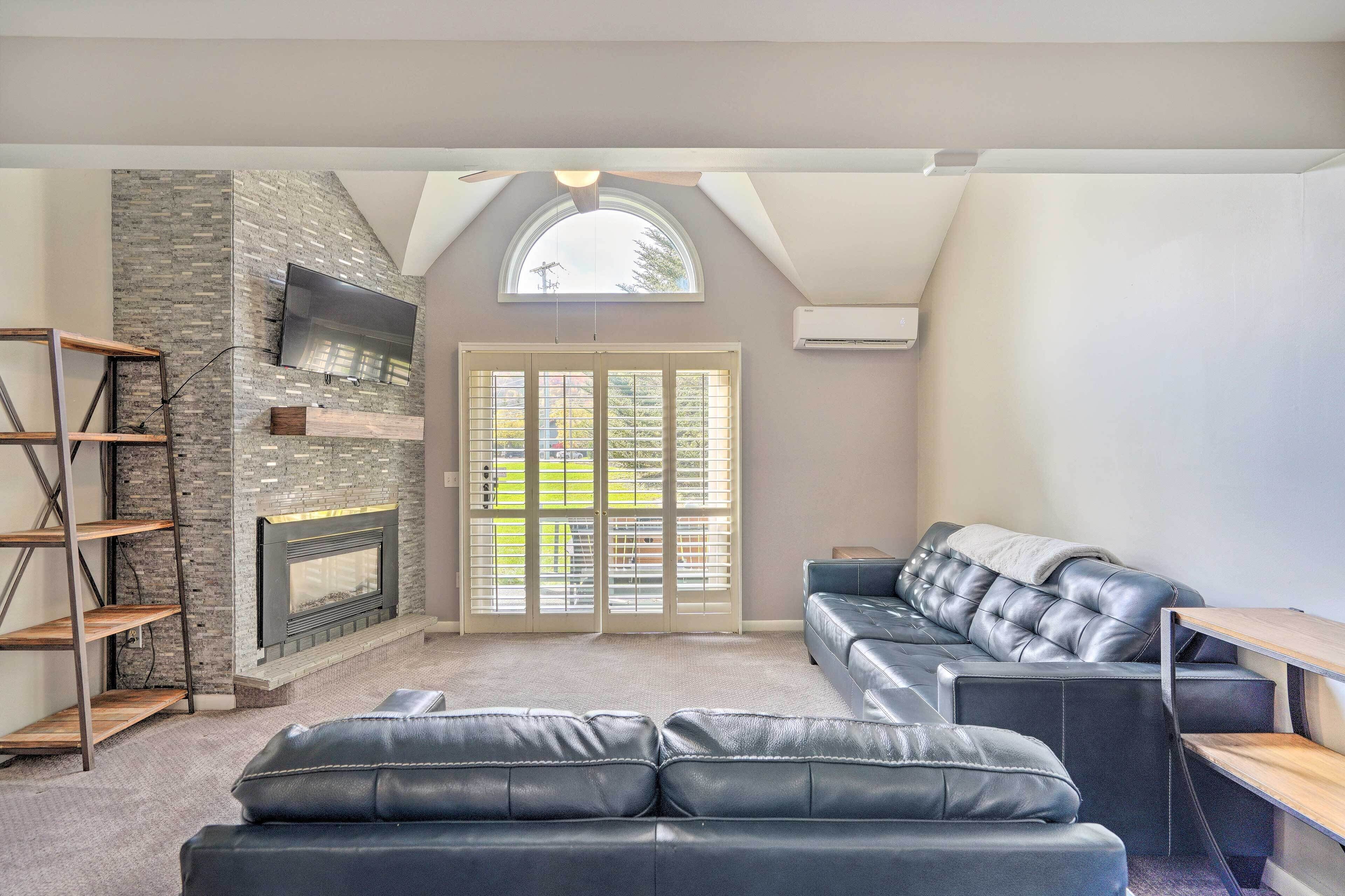 A modern living room with a fireplace, a black leather sofa, a wooden shelf, and a TV mounted on the wall.