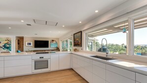 A modern kitchen with white cabinetry, a built-in oven, a large window with a view, and a flat-screen TV mounted above the counter. - Luxe Mid-century Austin Home w/ Canyon Views (Austin)