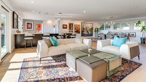 A spacious living area with a large sofa, a glass coffee table, and a patterned rug. There are multiple chairs and a dining area in the background. - Luxe Mid-century Austin Home w/ Canyon Views (Austin)
