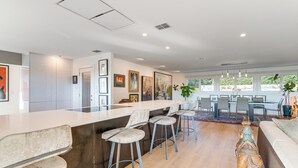 A modern kitchen with a bar area, wooden flooring, and a dining area with a large window. - Luxe Mid-century Austin Home w/ Canyon Views (Austin)