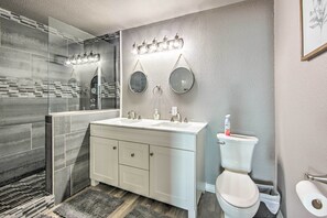 Ein modernes Badezimmer mit Duschkabine aus Glas, einem weißen Waschtisch mit zwei Waschbecken, einer Toilette und einer wandmontierten Leuchte.