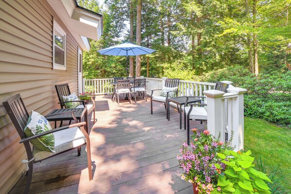 A wooden deck with outdoor furniture, including chairs and a table, surrounded by lush greenery.