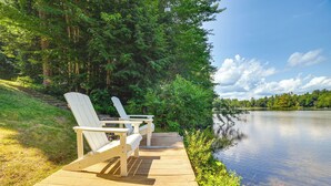 A wooden deck with two Adirondack chairs overlooking a calm lake, surrounded by lush greenery and trees. - Breathtaking Newbury Cottage w/ Deck on Chalk Pond (Newbury)