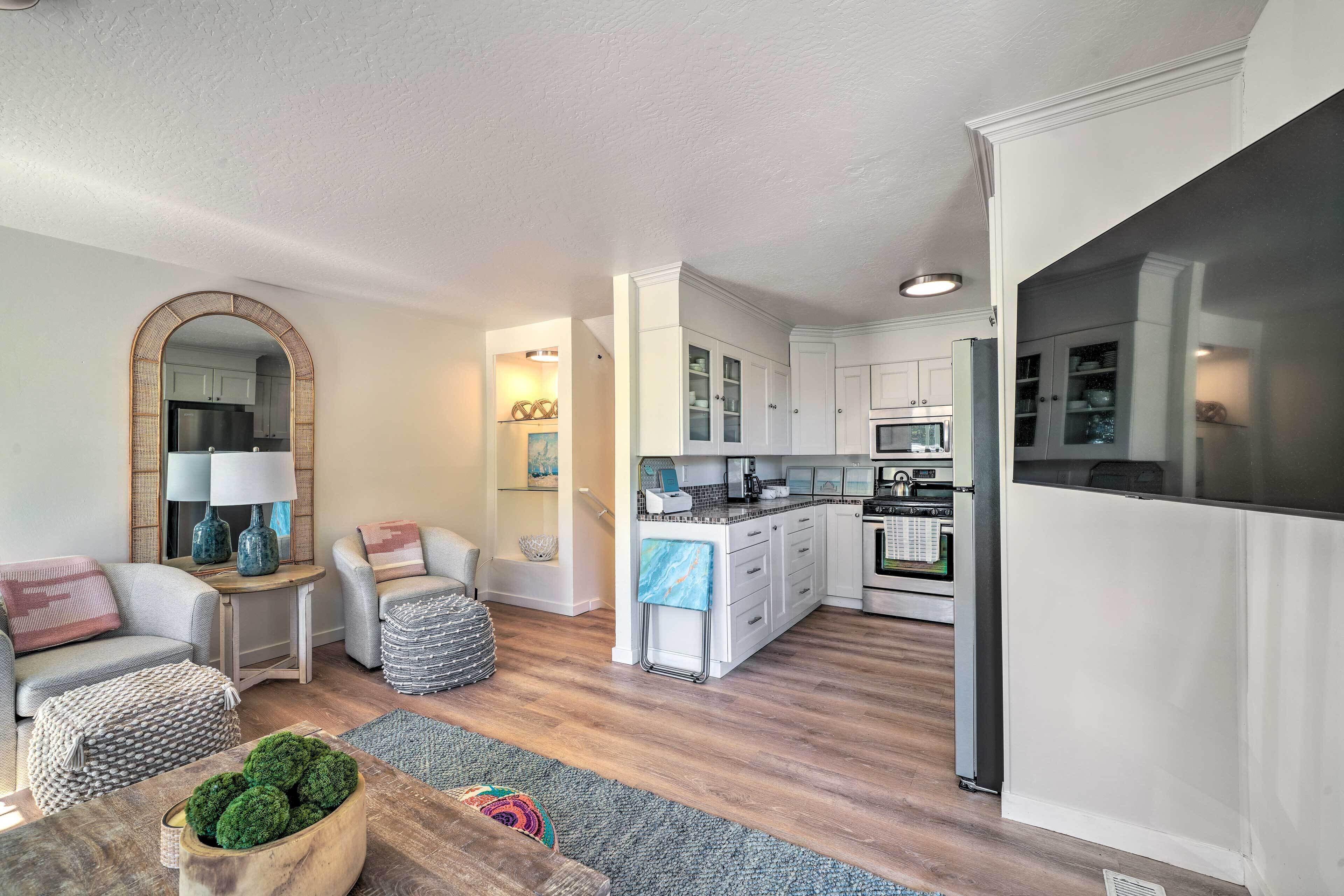 A modern kitchen with white cabinets, a refrigerator, and a microwave. There is a dining area with a table and chairs, and a living room with a sofa and a lamp.