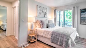 A bedroom with a bed, bedside lamps, a small wooden nightstand, and a window with a view of greenery.