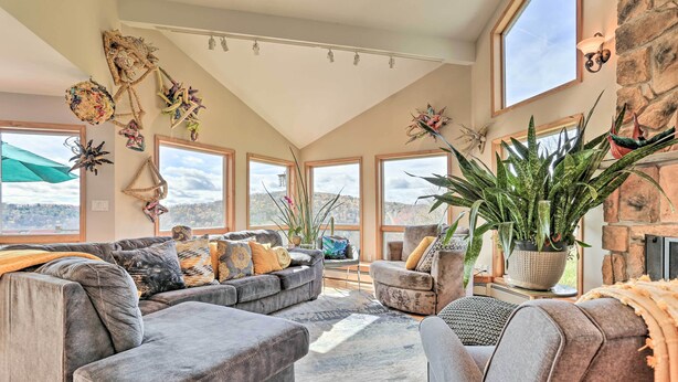 A spacious living room with a gray sectional sofa, a coffee table, and a fireplace. There are large windows offering a view of the outdoors.