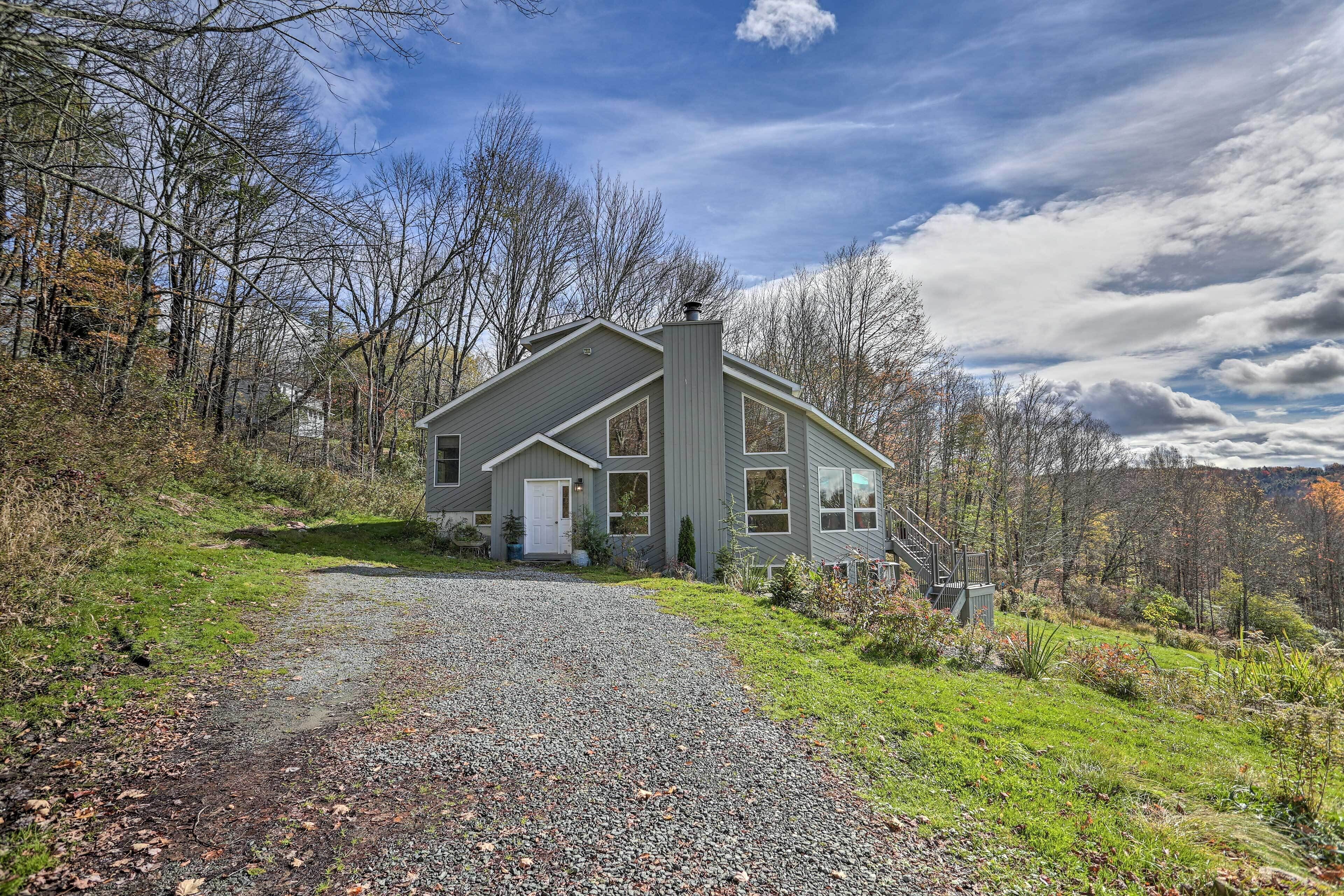 A two-story house with a gravel driveway leading to a white door, surrounded by trees and a clear sky.