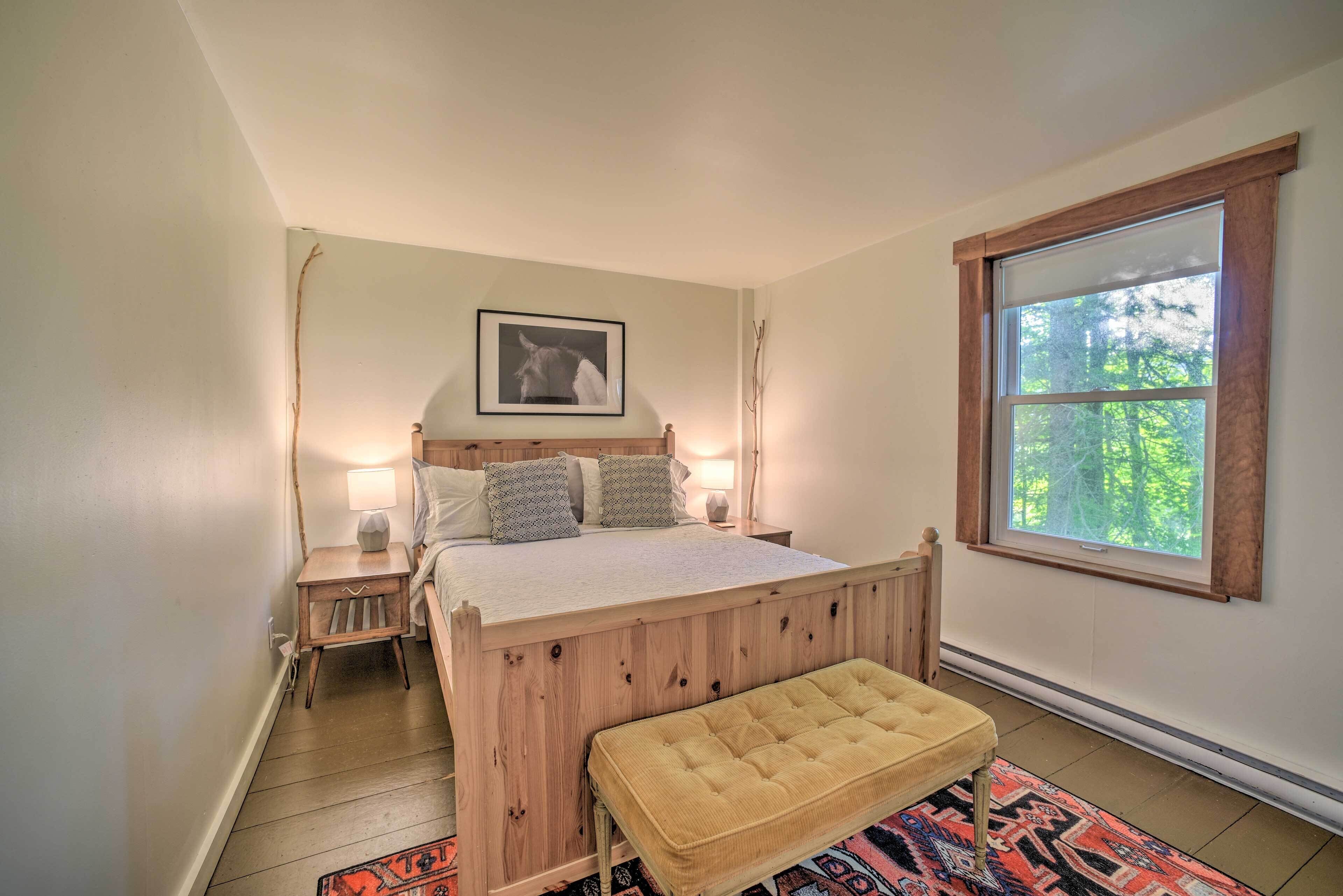 A bedroom with a wooden bed, bedside lamps, a bench, a rug, and a window with a view of trees.