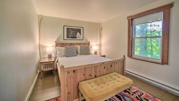 A bedroom with a wooden bed, bedside lamps, a bench, a rug, and a window with a view of trees.